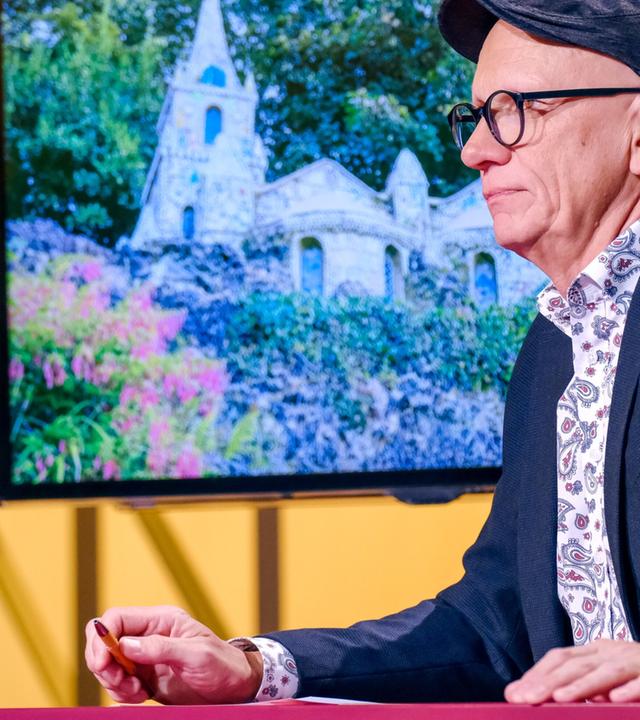 Das Bild zeigt einen Mann, der an einem Tisch sitzt und nach rechts schaut. Er trägt eine Schirmmütze und eine Brille. Sein Outfit besteht aus einem dunklen Sakko und einem bunteren Hemd mit einem Paisleymuster. Auf dem Tisch vor ihm steht ein Glas Wasser, und seine Hand hält einen Stift. Im Hintergrund ist ein Bildschirm zu sehen, der ein Bild eines architektonischen Bauwerks zeigt, das von üppigem Grün umgeben ist. Das Bauwerk hat spitze Türme und eine steinerne Fassade. Im Hintergrund ist eine weitere Person unscharf zu sehen, die anscheinend auf einem anderen Stuhl sitzt. Das gesamte Setting wirkt professionell und ist lebhaft beleuchtet.
