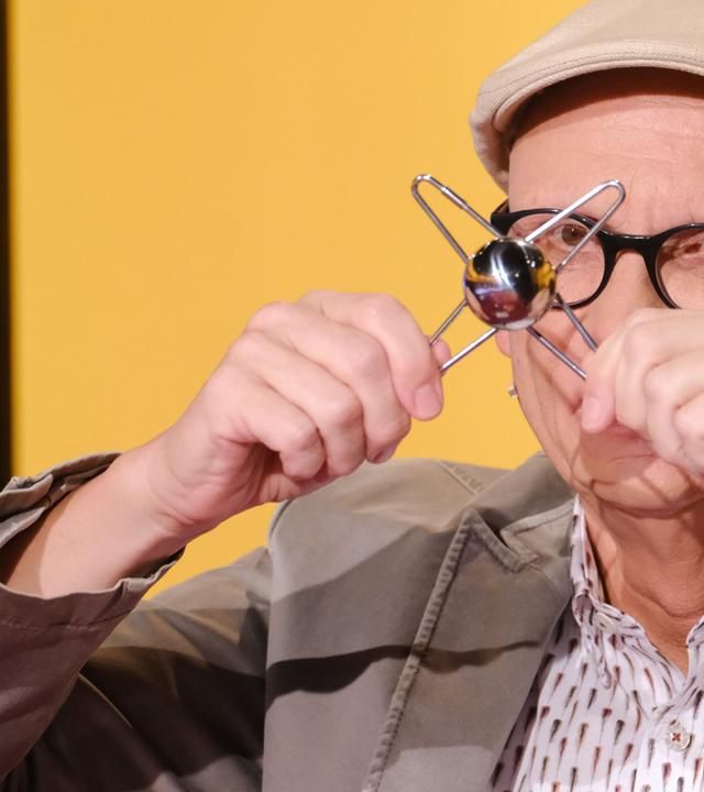 Das Bild zeigt einen Mann, der ein Werkzeug oder Spielzeug in den Händen hält, das aus einer runden Kugel und drei daran befestigten Metallarmen besteht. Der Mann trägt eine graue Jacke und ein Hemd mit einem Muster aus vielen kleinen, senkrechten Strichen. Er hat eine Glatze und trägt eine helle, runde Mütze sowie eine Brille mit dunklem Rahmen. Der Hintergrund ist gelb, und ein Teil des Bildes zeigt einen Bildschirm mit bunten Grafiken. Der Mann scheint humorvoll oder neugierig zu sein und richtet seine Aufmerksamkeit auf das Objekt in seinen Händen.