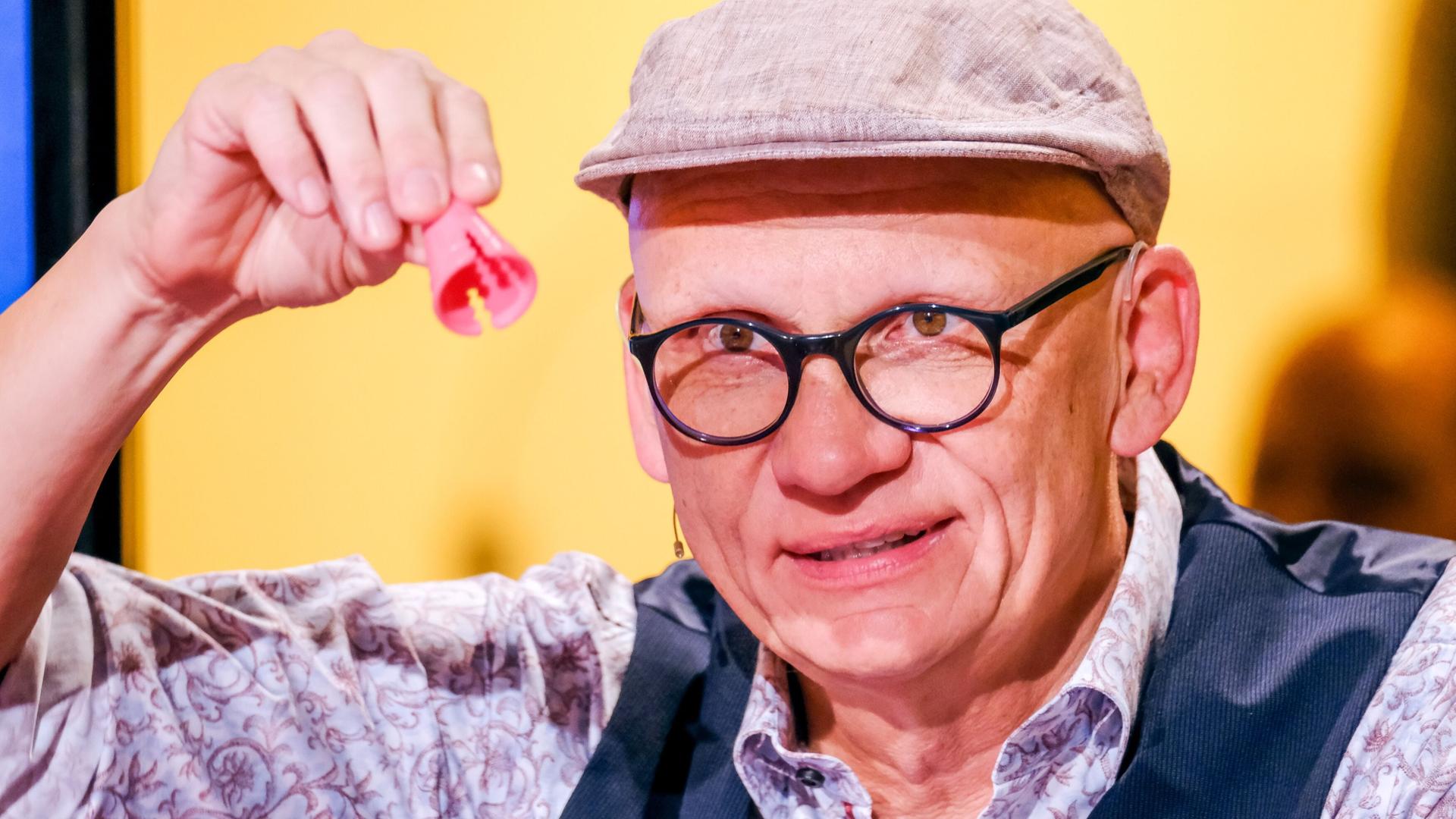 Auf dem Bild ist ein Mann zu sehen, der eine rote, klobige Figur mit zwei Öffnungen in der Hand hält. Er trägt eine graue Schirmmütze und eine Brille mit dünnem, schwarzem Gestell. Sein Gesicht hat ein freundliches und humorvolles Ausdruck. Er ist in einem gestreiften Hemd mit einem floralen Muster und einem dunklen Weste gekleidet. Der Hintergrund ist gelb und scheint aus einer Studio-Umgebung mit Beleuchtung zu stammen.