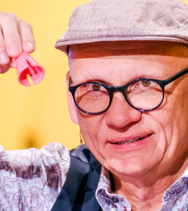 Auf dem Bild ist ein Mann zu sehen, der eine rote, klobige Figur mit zwei Öffnungen in der Hand hält. Er trägt eine graue Schirmmütze und eine Brille mit dünnem, schwarzem Gestell. Sein Gesicht hat ein freundliches und humorvolles Ausdruck. Er ist in einem gestreiften Hemd mit einem floralen Muster und einem dunklen Weste gekleidet. Der Hintergrund ist gelb und scheint aus einer Studio-Umgebung mit Beleuchtung zu stammen.