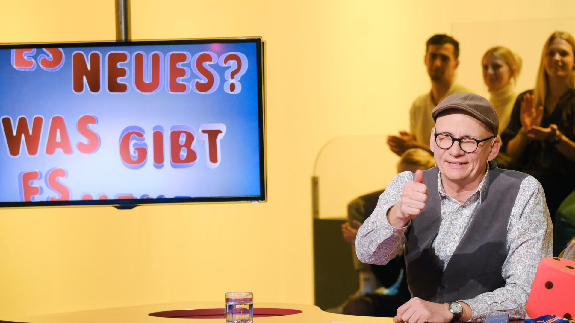 Das Bild zeigt eine Fernsehszene aus der Sendung "Was gibt es Neues?". Im Vordergrund sitzt ein Mann, der Oliver Baier heißt. Er trägt eine graue Kappe, eine Brille mit dunklem Rahmen und hat ein helles, gemustertes Hemd an, über dem er eine dunkle Weste trägt. Er lächelt und gibt mit der rechten Hand ein Daumenhoch. Vor ihm befindet sich ein runder Tisch, auf dem ein Glas mit Wasser steht.   Im Hintergrund ist ein Bildschirm zu sehen, auf dem der Text "ES NEUES? WAS GIBT ES NEUES?" in bunten Buchstaben dargestellt ist. Die Farbgebung im Hintergrund ist überwiegend gelb, und in der Nähe des Tisches sind mehrere Personen zu erkennen, die im Publikum sitzen und applaudieren.