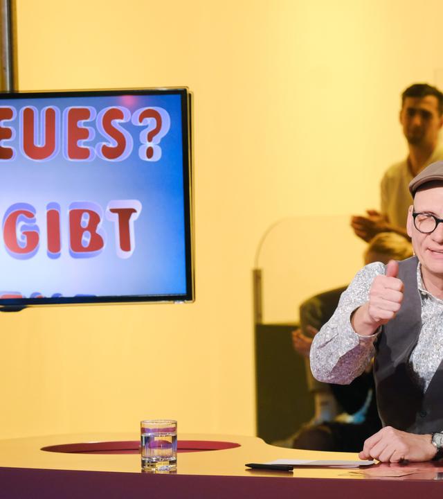 Das Bild zeigt eine Fernsehszene aus der Sendung "Was gibt es Neues?". Im Vordergrund sitzt ein Mann, der Oliver Baier heißt. Er trägt eine graue Kappe, eine Brille mit dunklem Rahmen und hat ein helles, gemustertes Hemd an, über dem er eine dunkle Weste trägt. Er lächelt und gibt mit der rechten Hand ein Daumenhoch. Vor ihm befindet sich ein runder Tisch, auf dem ein Glas mit Wasser steht.   Im Hintergrund ist ein Bildschirm zu sehen, auf dem der Text "ES NEUES? WAS GIBT ES NEUES?" in bunten Buchstaben dargestellt ist. Die Farbgebung im Hintergrund ist überwiegend gelb, und in der Nähe des Tisches sind mehrere Personen zu erkennen, die im Publikum sitzen und applaudieren.