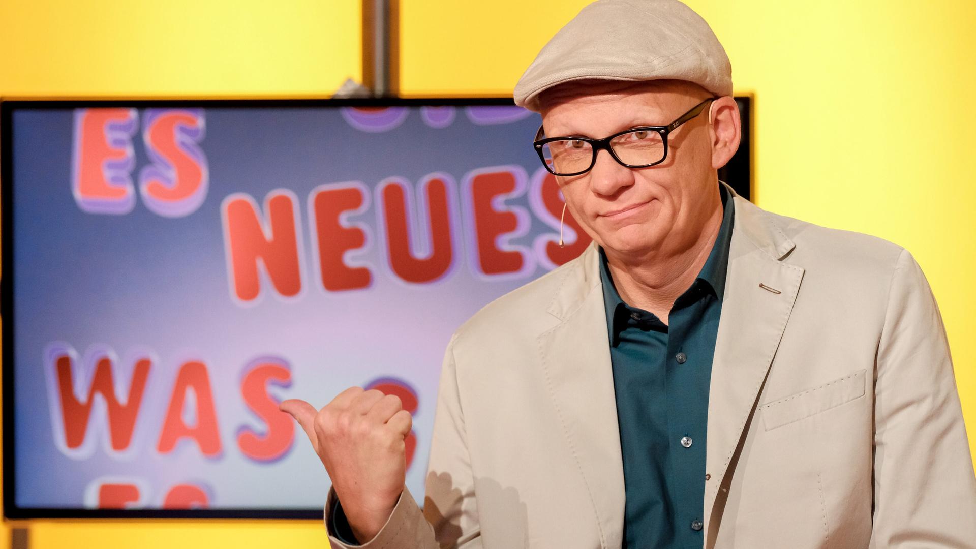 Das Bild zeigt einen Mann, der an einem Tisch sitzt. Er trägt eine graue Jacke, ein weißes Hemd mit einem blau-weißen Muster und eine graue Schirmmütze. Vor ihm steht ein Glas Wasser und er hält einen Stift in der Hand. Im Hintergrund ist ein großer Bildschirm zu sehen, auf dem ein Tier, vermutlich ein Ameisenigel, abgebildet ist. Der Hintergrund des Raumes ist gelb, was eine warmes Lichtambiente schafft. Die Szene scheint aus einer Fernsehsendung zu stammen.
