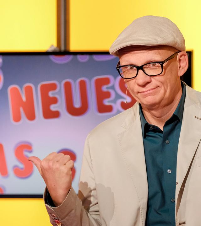 Das Bild zeigt einen Mann, der an einem Tisch sitzt. Er trägt eine graue Jacke, ein weißes Hemd mit einem blau-weißen Muster und eine graue Schirmmütze. Vor ihm steht ein Glas Wasser und er hält einen Stift in der Hand. Im Hintergrund ist ein großer Bildschirm zu sehen, auf dem ein Tier, vermutlich ein Ameisenigel, abgebildet ist. Der Hintergrund des Raumes ist gelb, was eine warmes Lichtambiente schafft. Die Szene scheint aus einer Fernsehsendung zu stammen.