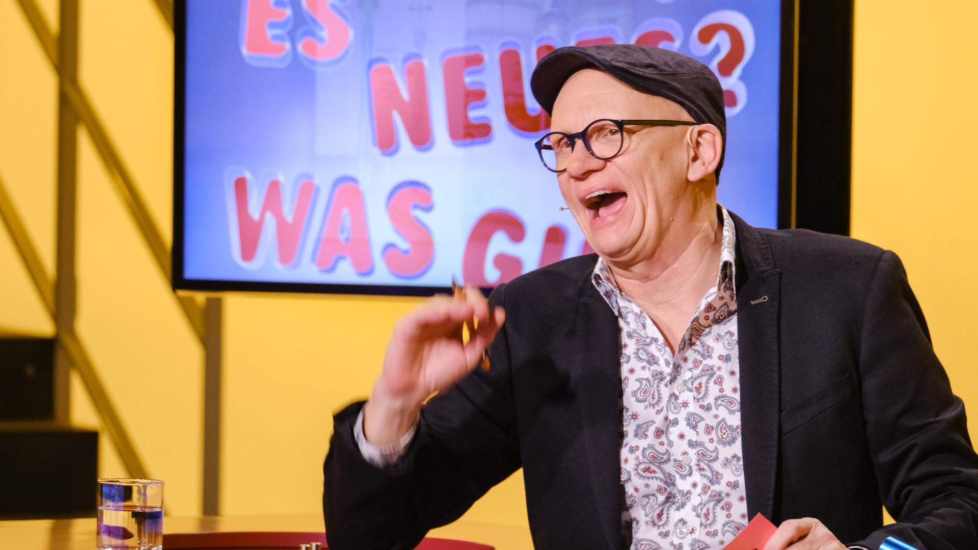 Das Bild zeigt eine Szene aus einer Fernsehsendung mit einem Moderator, der in einem Studio sitzt. Der Moderator hat eine Glatze, trägt eine Brille und eine graue Mütze. Er ist lachend abgebildet und hält ein rotes Blatt Papier in der Hand. Hinter ihm ist ein Bildschirm zu sehen, auf dem in großen, bunten Buchstaben steht: "ES GIBT NEUES? WAS GIBT?" Die Hintergrundfarbe des Bildschirms ist blau und gelb. Der Tisch, an dem der Moderator sitzt, hat eine Holzoberfläche mit runden roten Einlagen. Auf dem Tisch steht ein Wasserglas. Die Szene vermittelt eine fröhliche und unterhaltsame Atmosphäre.