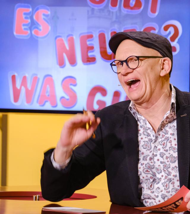 Das Bild zeigt eine Szene aus einer Fernsehsendung mit einem Moderator, der in einem Studio sitzt. Der Moderator hat eine Glatze, trägt eine Brille und eine graue Mütze. Er ist lachend abgebildet und hält ein rotes Blatt Papier in der Hand. Hinter ihm ist ein Bildschirm zu sehen, auf dem in großen, bunten Buchstaben steht: "ES GIBT NEUES? WAS GIBT?" Die Hintergrundfarbe des Bildschirms ist blau und gelb. Der Tisch, an dem der Moderator sitzt, hat eine Holzoberfläche mit runden roten Einlagen. Auf dem Tisch steht ein Wasserglas. Die Szene vermittelt eine fröhliche und unterhaltsame Atmosphäre.
