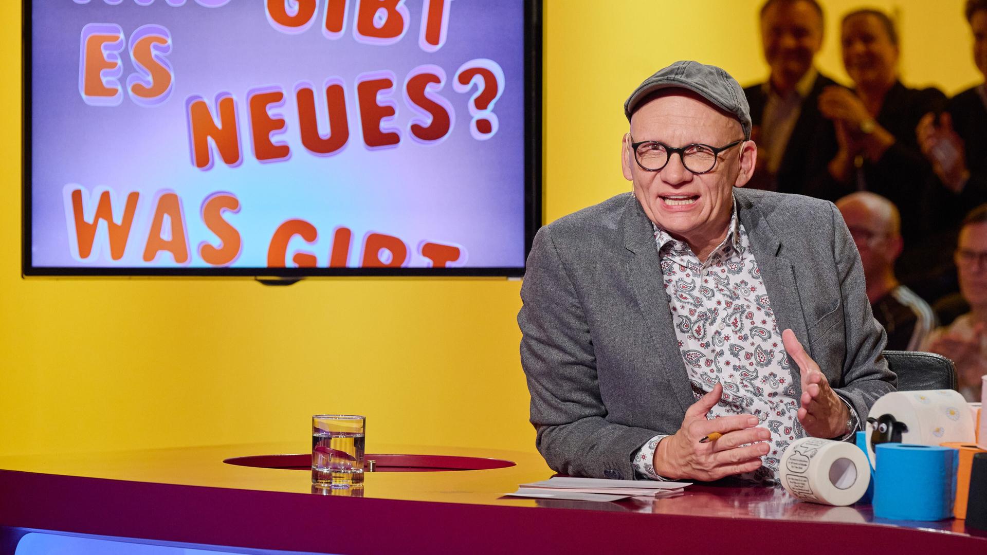 Auf dem Bild sieht man einen Raum mit einem hellen, gelben Hintergrund. Im Vordergrund befindet sich ein runder Tisch in den Farben Rot und Blau. An diesem Tisch sitzt ein Mann, der eine graue Jacke und ein bunt gemustertes Hemd trägt sowie eine graue Mütze. Er hat eine Brille auf und gestikuliert mit seinen Händen, während er spricht. Auf dem Tisch steht ein Glas mit Wasser.  Im Hintergrund ist ein Bildschirm zu sehen, auf dem in bunten Buchstaben die Wörter „Was gibt es Neues?“ zu lesen sind. Hinter dem Tisch sind einige Personen im Publikum sichtbar, die applaudieren oder aufmerksam dem Geschehen folgen. Die gesamte Szene vermittelt eine fröhliche und unterhaltsame Atmosphäre.