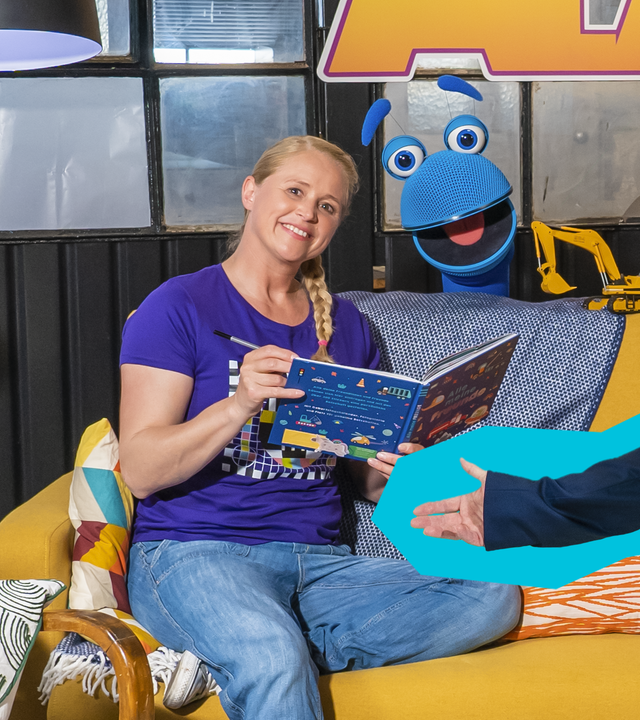 Singa sitzt lächelnd mit einem Buch in der Hand auf dem Sofa. Das SingDing schaut lachend hinter dem Sofa hervor. Volker Rosin steht mit ausgebreiteten Armen und einer Lokführermütze vor dem Sofa und lächelt ebenfalls in die Kamera.