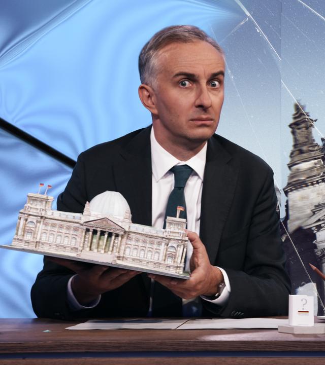 Jan Böhmermann sitzt am Schreibtisch und hält ein Modell des Reichstagsgebäudes in den Händen. Im Hintergrund ist ein visuell verzerrtes Bild des Reichstagsgebäudes zu sehen. 