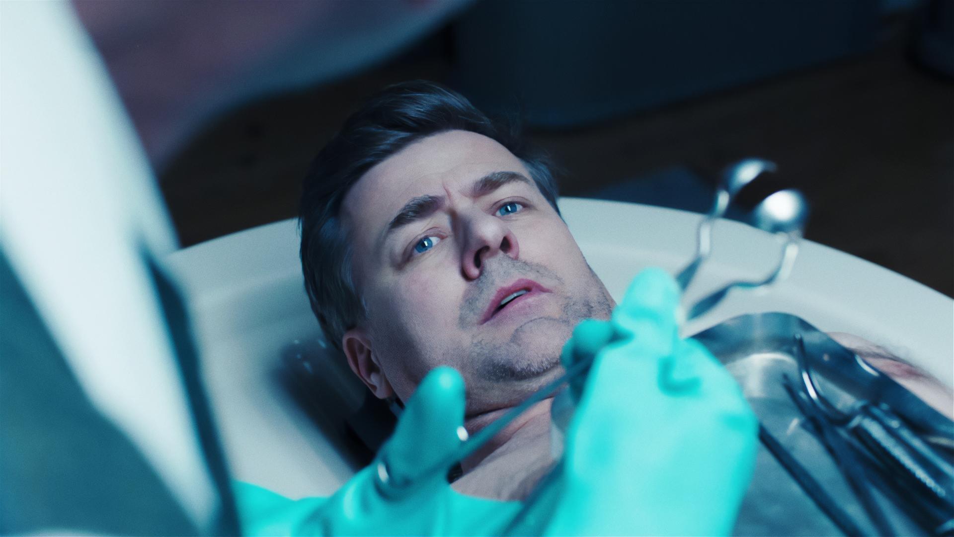 Der besorgt aussehende Harald Neuhauser (Marcus Mittermeier) liegt oberkörperfrei auf einem Obduktionstisch. Eine andere Person in OP-Kleidung, von der nur ein Teil des Körpers zu sehen ist, steht über ihm und hält in den behandschuhten Händen eine Zange.