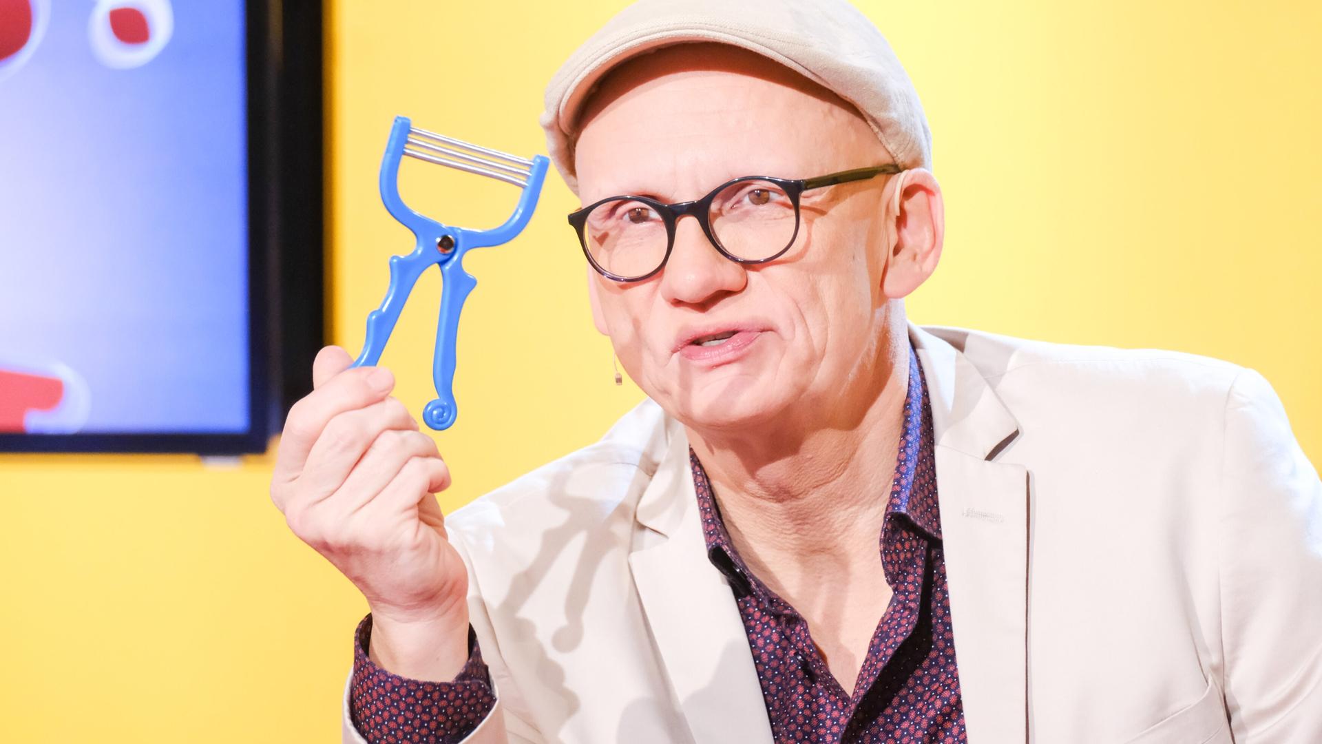 Das Bild zeigt einen Mann, der einen blauen Gemüseschäler in der Hand hält. Er trägt eine helle Jacke und eine Kappe. Der Mann hat eine Brille auf und steht vor einem Hintergrund in Gelb, der mit grafischen Elementen gestaltet ist. Unmittelbar hinter ihm ist ein Bildschirm sichtbar, auf dem teilweise farbige Fragezeichen zu sehen sind. Der Ausdruck des Mannes wirkt konzentriert und er spricht offensichtlich in eine Kamera.