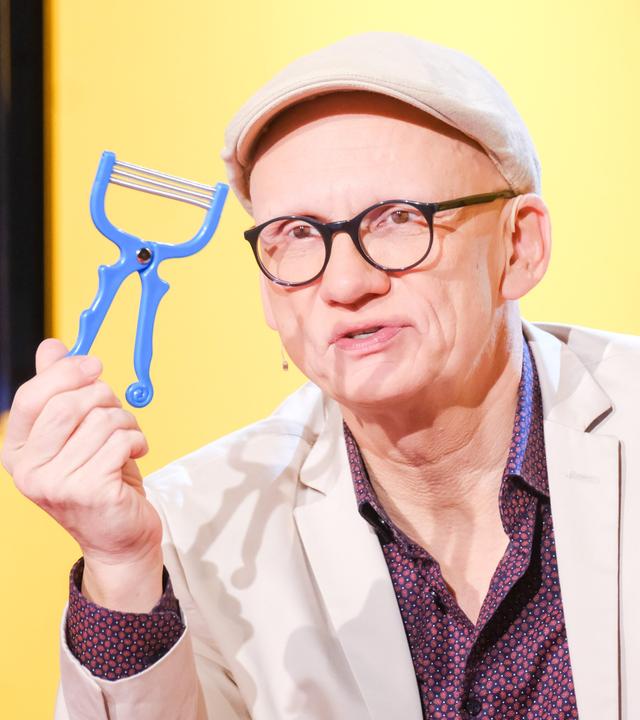 Das Bild zeigt einen Mann, der einen blauen Gemüseschäler in der Hand hält. Er trägt eine helle Jacke und eine Kappe. Der Mann hat eine Brille auf und steht vor einem Hintergrund in Gelb, der mit grafischen Elementen gestaltet ist. Unmittelbar hinter ihm ist ein Bildschirm sichtbar, auf dem teilweise farbige Fragezeichen zu sehen sind. Der Ausdruck des Mannes wirkt konzentriert und er spricht offensichtlich in eine Kamera.