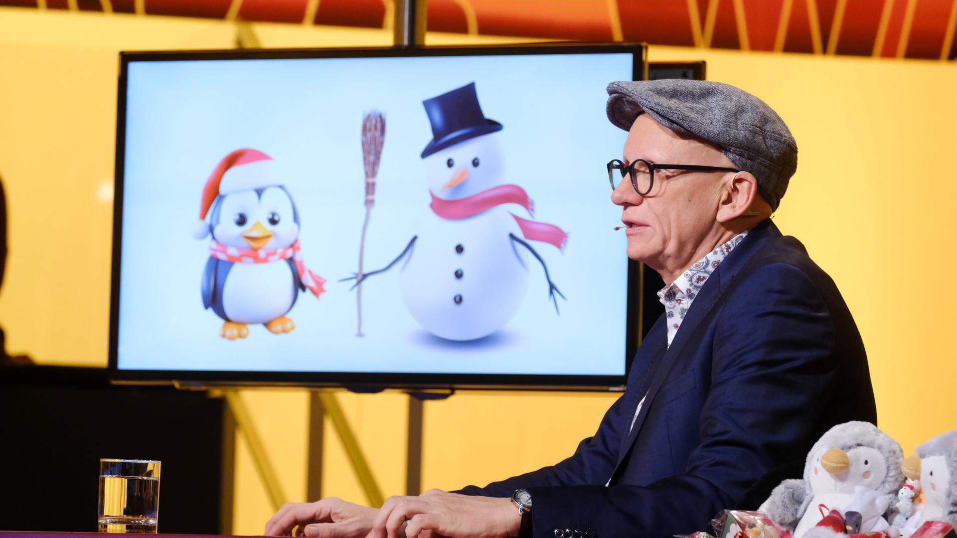 Das Bild zeigt einen männlichen Moderator mit einer schräg sitzenden Mütze und einer Brille. Er sitzt an einem Tisch, auf dem ein Glas Wasser steht. Im Hintergrund befindet sich ein Bildschirm, auf dem zwei cartoonhafte Figuren abgebildet sind: ein Pinguin mit einem roten Schal und einer Weihnachtsmütze sowie ein Schneemann mit einem schwarzen Zylinder und einem roten Schal. Der Tisch ist mit kleinen Dekorationsfiguren von Pinguinen und Schneemännern geschmückt. Der Hintergrund ist in warmen Gelbtönen gehalten, und das Bild vermittelt eine festliche Atmosphäre.