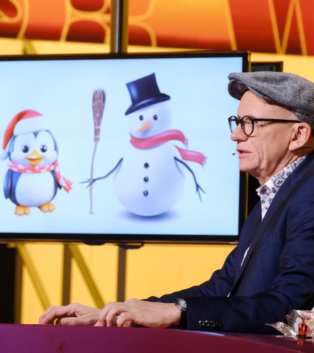 Das Bild zeigt einen männlichen Moderator mit einer schräg sitzenden Mütze und einer Brille. Er sitzt an einem Tisch, auf dem ein Glas Wasser steht. Im Hintergrund befindet sich ein Bildschirm, auf dem zwei cartoonhafte Figuren abgebildet sind: ein Pinguin mit einem roten Schal und einer Weihnachtsmütze sowie ein Schneemann mit einem schwarzen Zylinder und einem roten Schal. Der Tisch ist mit kleinen Dekorationsfiguren von Pinguinen und Schneemännern geschmückt. Der Hintergrund ist in warmen Gelbtönen gehalten, und das Bild vermittelt eine festliche Atmosphäre.