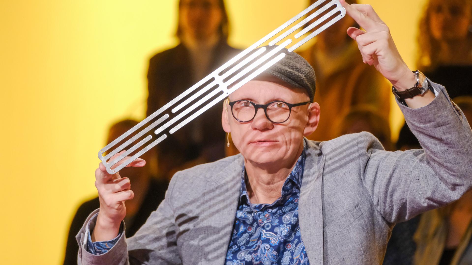 Das Bild zeigt einen Mann, der auf einem Tisch sitzt und eine lange, schmale Metallplatte in der Hand hält. Er trägt eine graue Jacke über einem blauen Hemd mit einem auffälligen Muster. Auf dem Kopf hat er eine graue Mütze. Der Mann blickt direkt in die Kamera und hat einen konzentrierten Ausdruck. Im Hintergrund sind unscharfe Silhouetten von Personen zu erkennen, die im Publikum sitzen. Die Beleuchtung ist sanft und hat einen gelben Farbton, was eine warme Atmosphäre schafft.