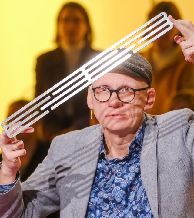 Das Bild zeigt einen Mann, der auf einem Tisch sitzt und eine lange, schmale Metallplatte in der Hand hält. Er trägt eine graue Jacke über einem blauen Hemd mit einem auffälligen Muster. Auf dem Kopf hat er eine graue Mütze. Der Mann blickt direkt in die Kamera und hat einen konzentrierten Ausdruck. Im Hintergrund sind unscharfe Silhouetten von Personen zu erkennen, die im Publikum sitzen. Die Beleuchtung ist sanft und hat einen gelben Farbton, was eine warme Atmosphäre schafft.