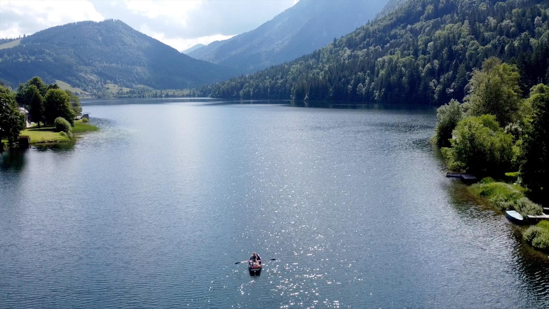 Ein idyllischer Bergsee liegt ruhig zwischen bewaldeten Hügeln, umgeben von Nebel und Felsen.