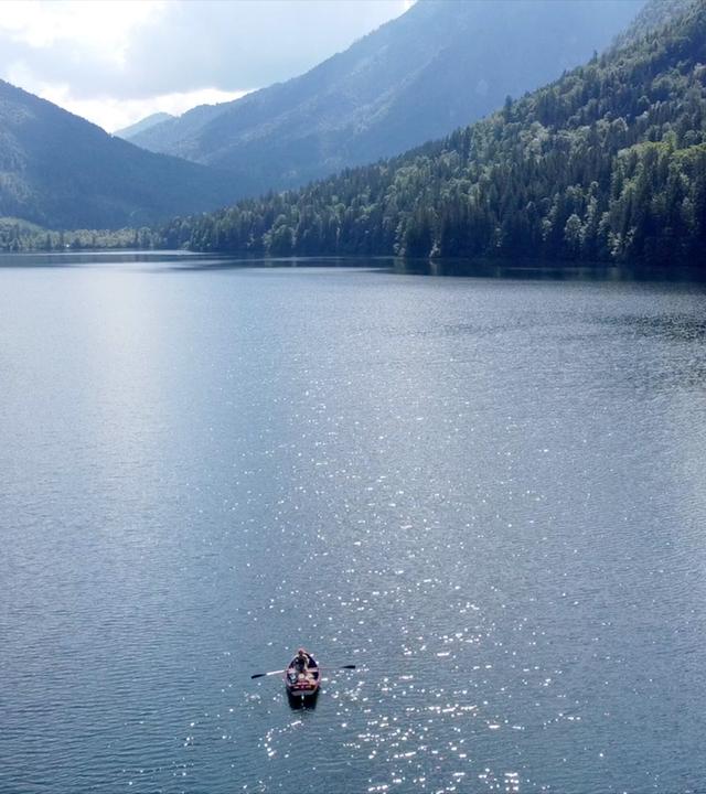 Ein idyllischer Bergsee liegt ruhig zwischen bewaldeten Hügeln, umgeben von Nebel und Felsen.