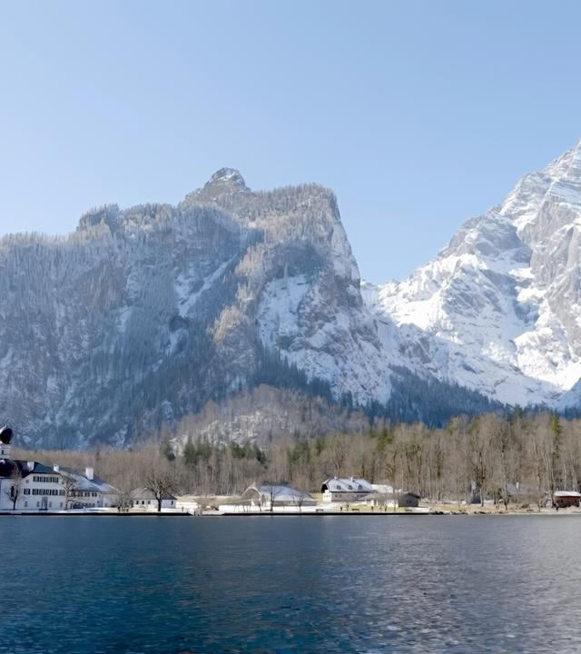 Das Bild zeigt eine winterliche Landschaft im Berchtesgadener Land, mit dem Königssee im Vordergrund und der markanten Ostwand des Watzmann im Hintergrund. Auf der linken Seite des Bildes ist die weithin bekannte Kirche St. Bartholomä zu sehen, eine auffällige, weiße Baufläche mit zwei charakteristischen, runden Türmen. Der See ist ruhig und spiegelnd, was die umliegenden verschneiten Berge reflektiert.  Im Hintergrund erkennt man große, steile Bergformationen mit schneebedeckten Gipfeln, die den Eindruck von majestätischer Erhabenheit erwecken. Die Bäume am Ufer sind kahl, während die Region von Schnee bedeckt ist, was eine ruhige und friedliche Atmosphäre schafft. Der Himmel ist klar und blau, was typisch für kalte Wintertage in den Alpen ist. Das Gesamtbild vermittelt ein Gefühl von natürlicher Schönheit und Klarheit in einer beeindruckenden Berglandschaft.