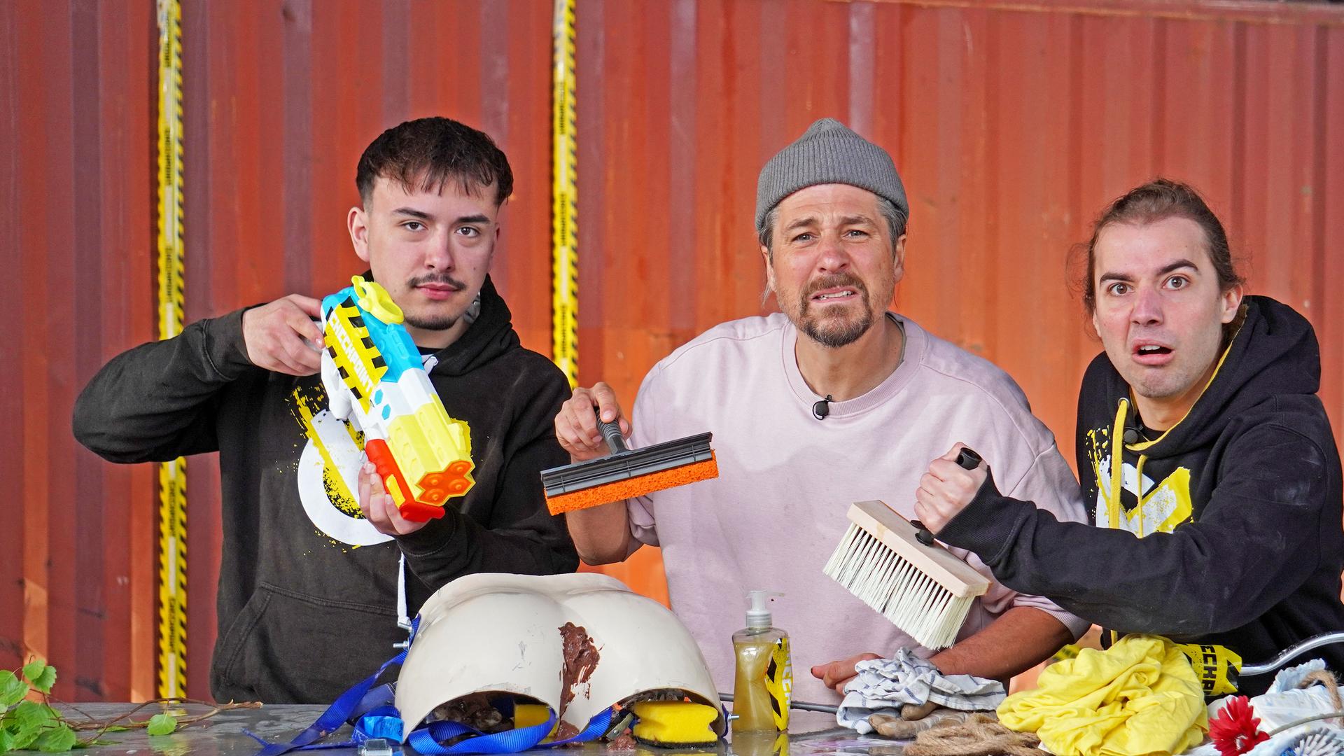 Moritz, Tommy Scheel und Leopold halten eine Wasserpistole, einen Fensterreiniger und eine Kleisterbürste in der Hand. Vor ihnen liegen diverse Utensilien auf dem Tisch.
