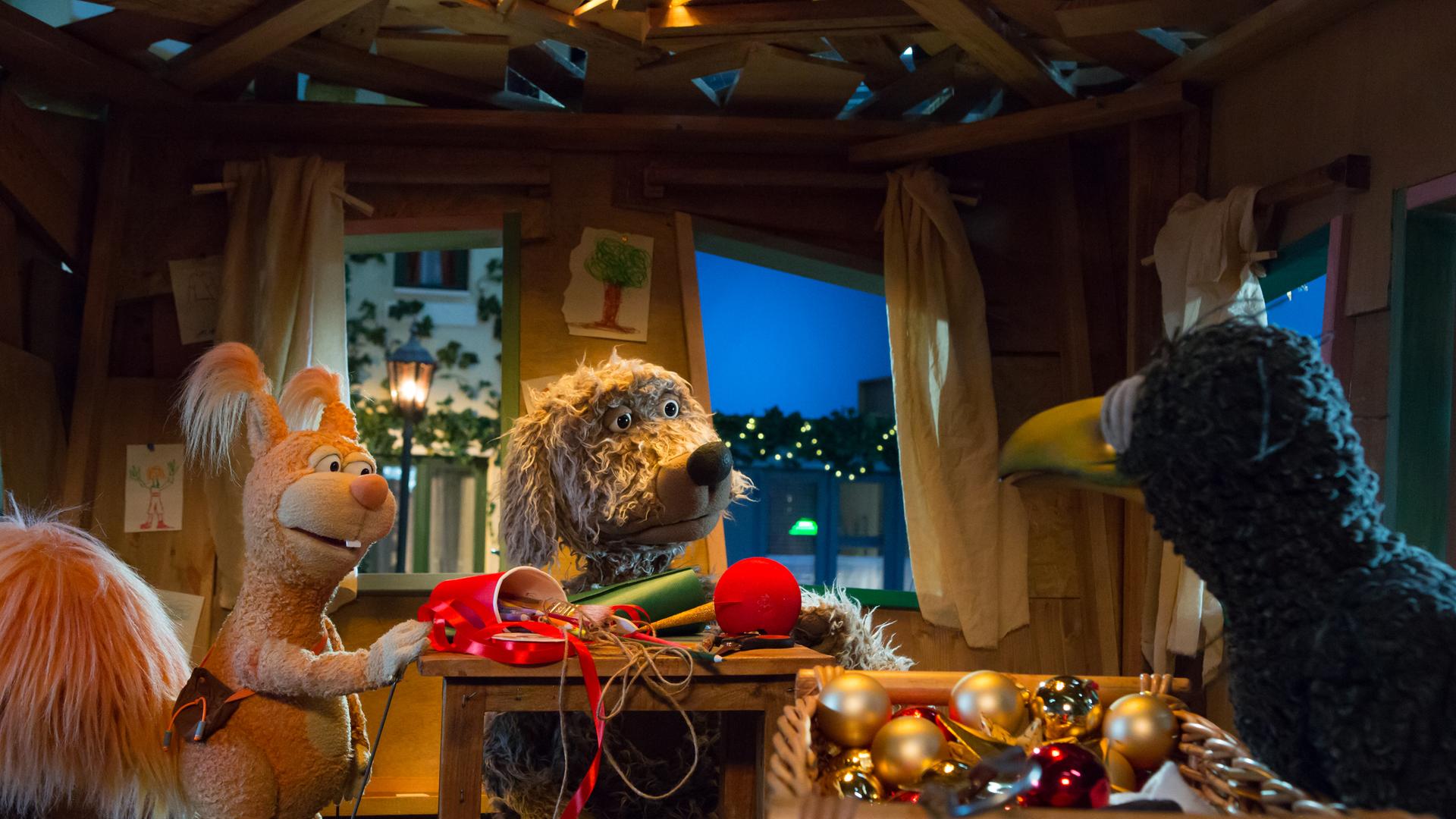 Ella, Buffo und Rudi sind im Baumhaus und schauen sich an. Zwischen ihnen auf dem Tisch liegen Bastelutensilien, im Vordergrund Weihnachtsbaumkugeln.