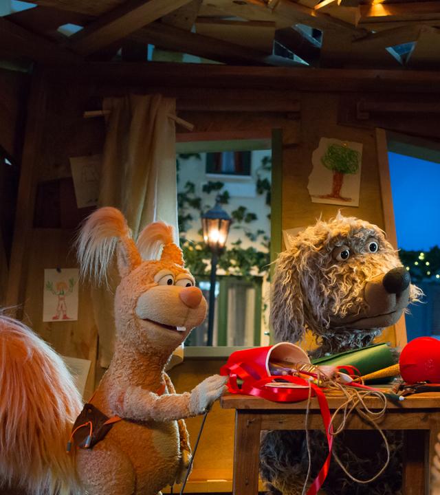 Ella, Buffo und Rudi sind im Baumhaus und schauen sich an. Zwischen ihnen auf dem Tisch liegen Bastelutensilien, im Vordergrund Weihnachtsbaumkugeln.