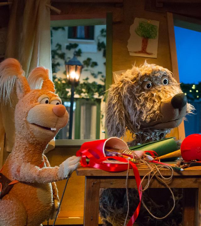 Ella, Buffo und Rudi sind im Baumhaus und schauen sich an. Zwischen ihnen auf dem Tisch liegen Bastelutensilien, im Vordergrund Weihnachtsbaumkugeln.