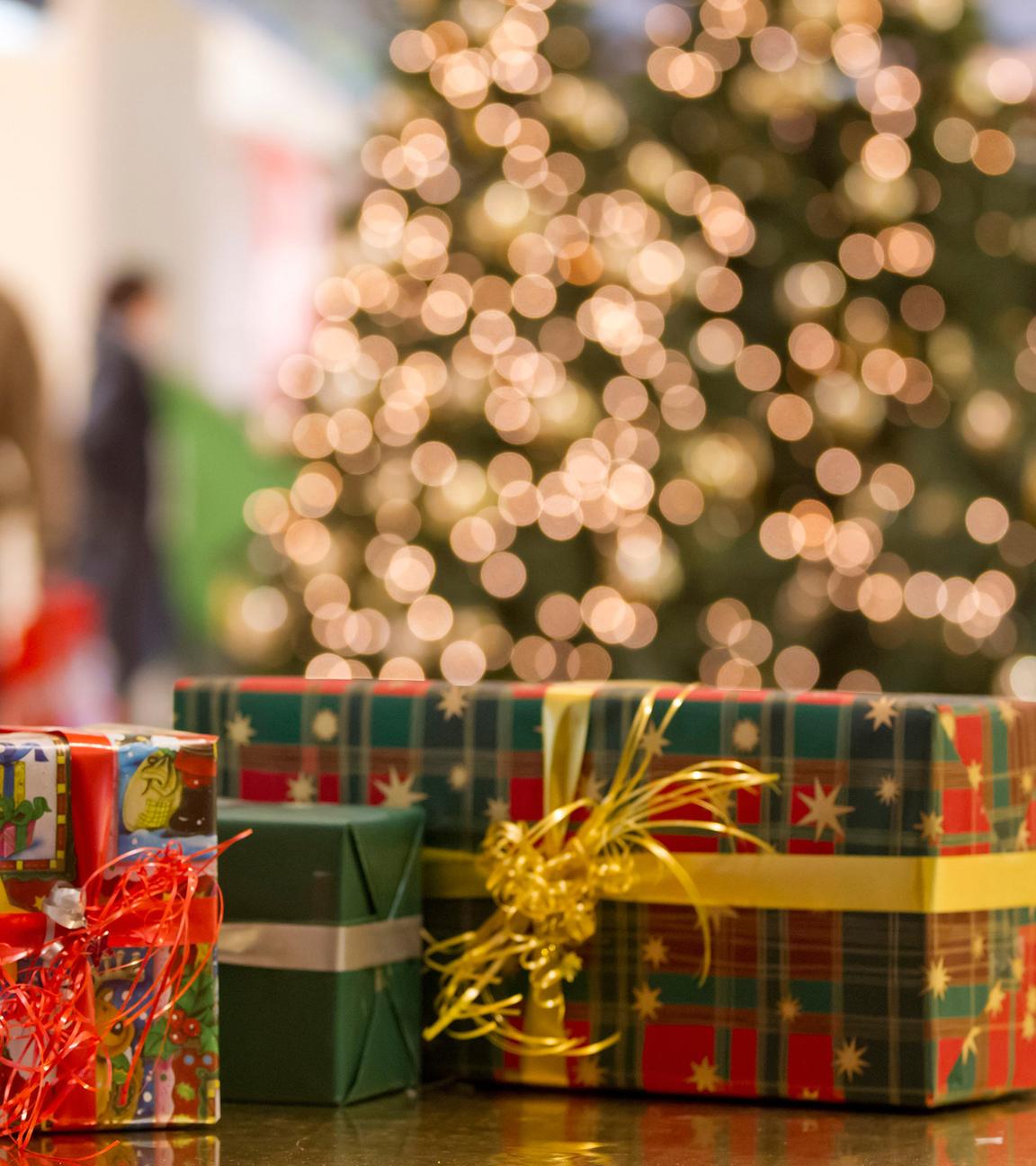 Vier Weihnachtsgeschenke liegen nebeneinander, im Hintergrund steht ein geschmückter Weihnachtsbaum und Menschen gehen vorbei.