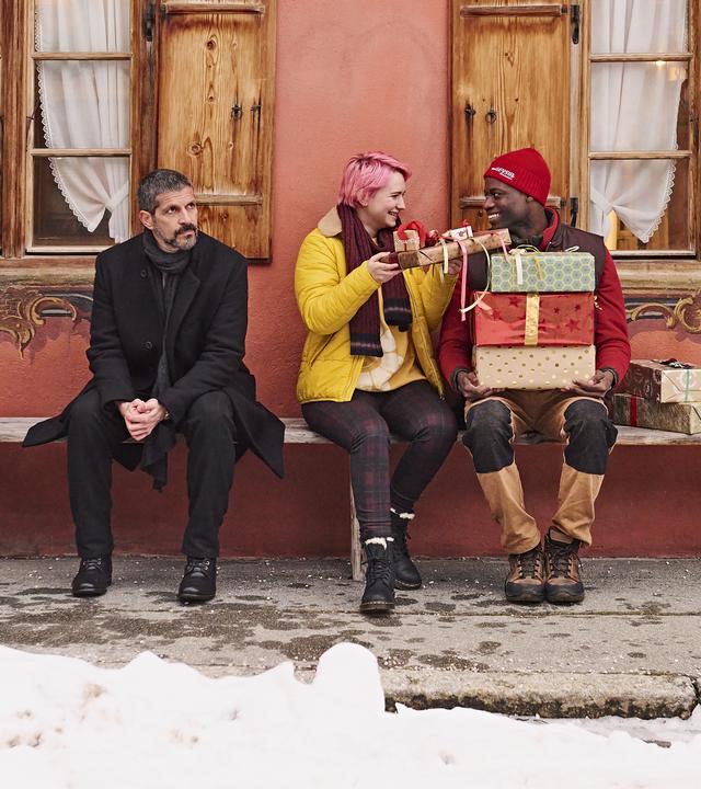 Adika sitzt auf einer Bank mit mehreren Geschenken auf dem Schoß. Tessa reicht ihm ein kleines Geschenk. Neben ihnen sitzt eine weitere Person. Links steht ein geschmückter Weihnachtsbaum, Schnee liegt am Boden.