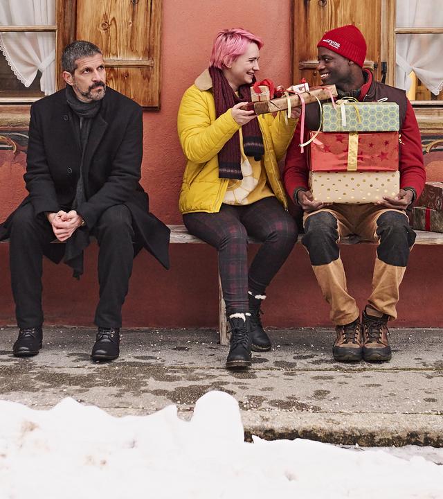 Adika sitzt auf einer Bank mit mehreren Geschenken auf dem Schoß. Tessa reicht ihm ein kleines Geschenk. Neben ihnen sitzt eine weitere Person. Links steht ein geschmückter Weihnachtsbaum, Schnee liegt am Boden.