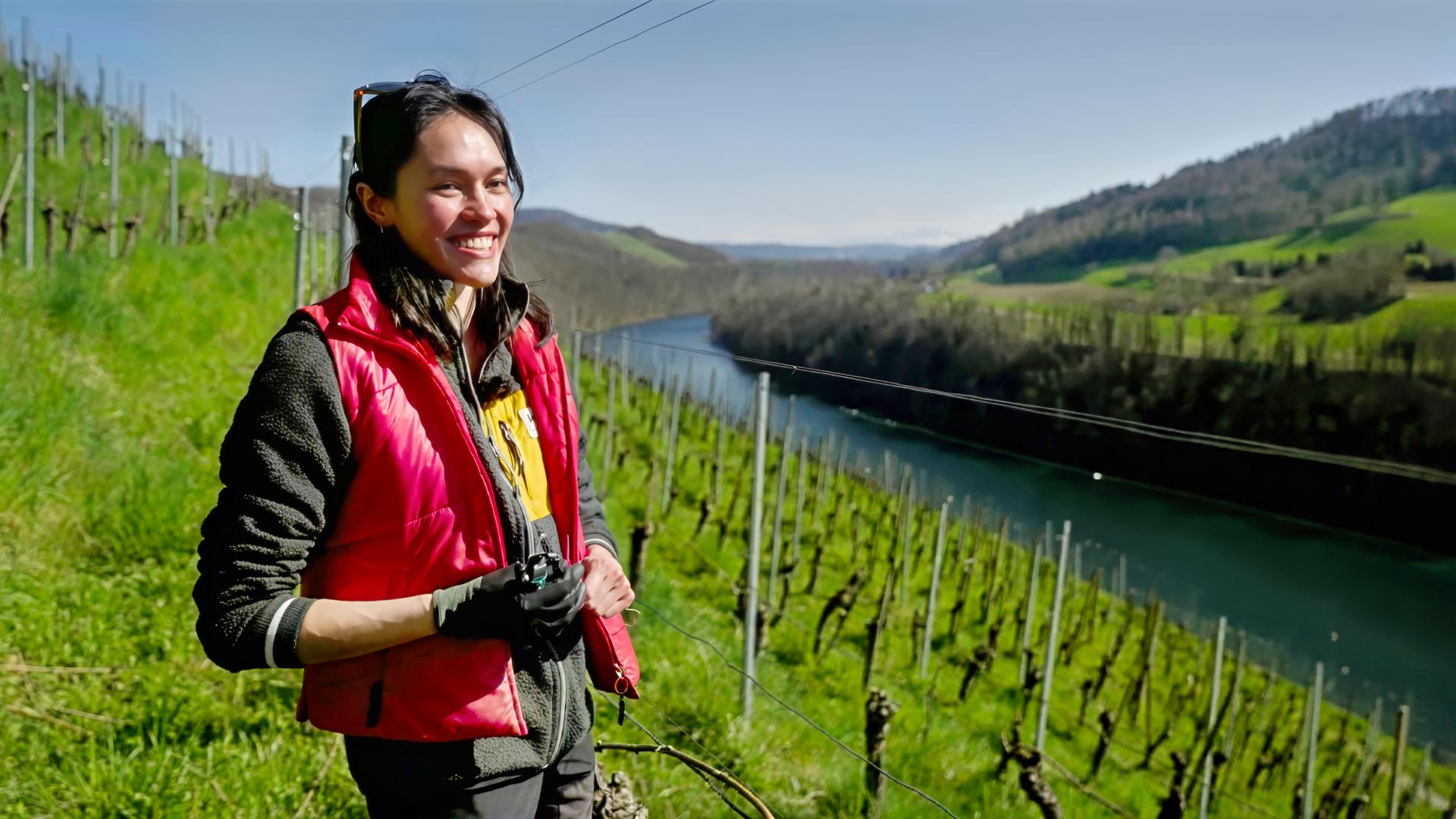 Junge Frau mit roter Weste steht auf einem sonnigen Weinberg oberhalb eines Flusses, im Hintergrund grüne Hügel und Rebenreihen.