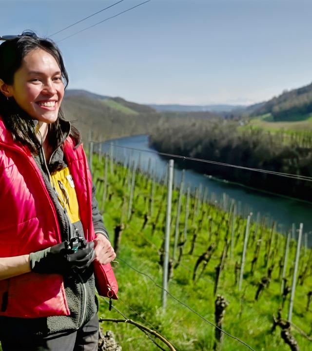 Junge Frau mit roter Weste steht auf einem sonnigen Weinberg oberhalb eines Flusses, im Hintergrund grüne Hügel und Rebenreihen.
