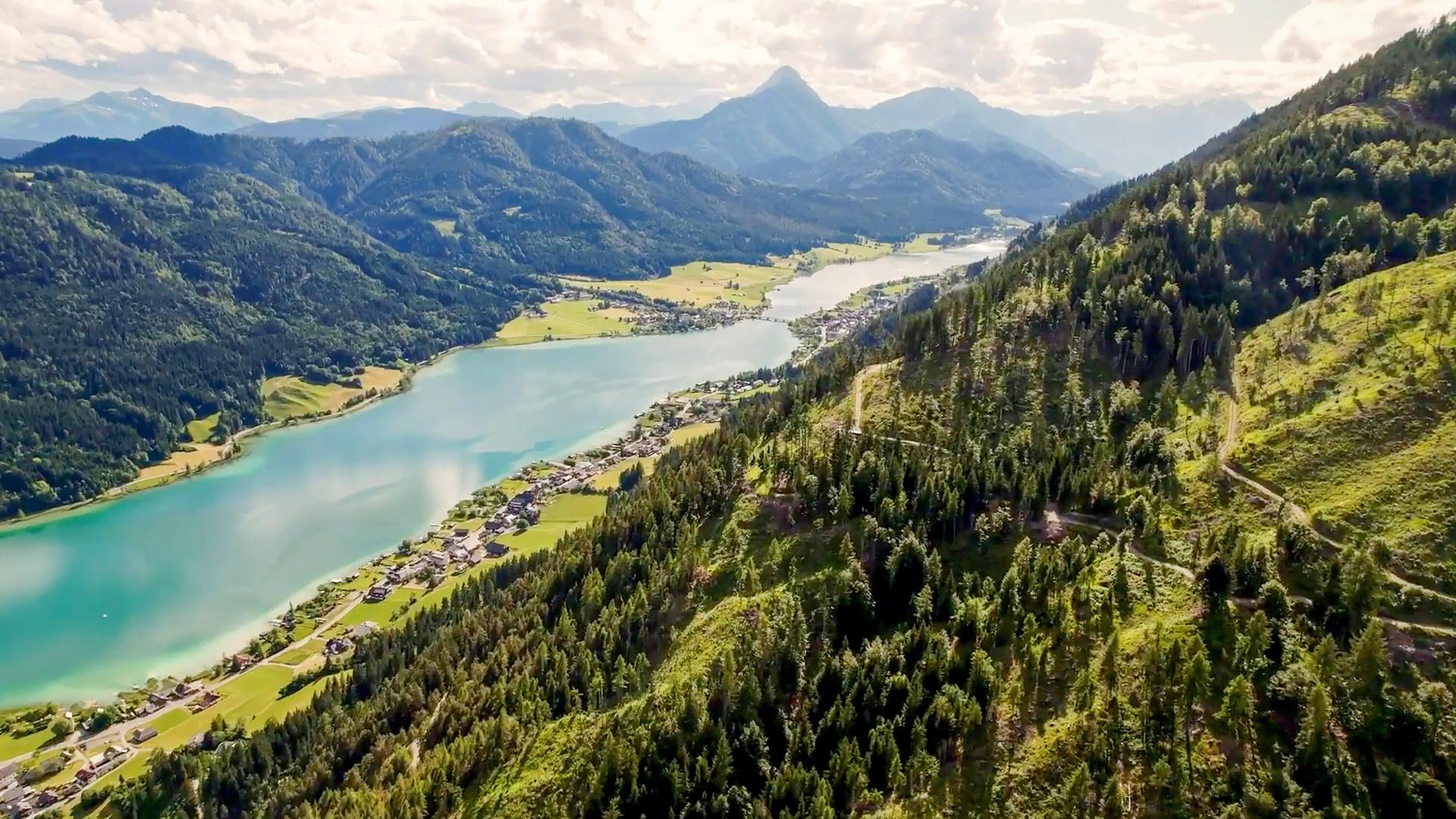 Das Bild zeigt eine panoramaartige Landschaft des Weißensees in den österreichischen Alpen. Der See hat eine tiefblaue Farbe und wird von angrenzenden Bergen und Wäldern umrahmt. Auf der linken Seite ist eine kleine Ansiedlung mit mehreren Gebäuden zu erkennen, die sich entlang des Ufers erstreckt. Die Umgebung ist mit grünen Wiesen und Bäumen bewachsen. Im Hintergrund erheben sich die hohen, schroffen Berge der Karnischen Alpen, die mit einigen bewaldeten Hängen und schneebedeckten Gipfeln versehen sind. Die Himmel ist teilweise bewölkt, was für eine weiche Lichtstimmung sorgt. Die gesamte Szene vermittelt ein Gefühl von Ruhe und natürlicher Schönheit.