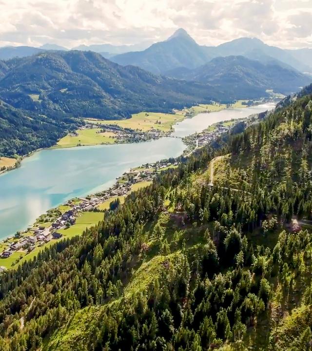 Das Bild zeigt eine panoramaartige Landschaft des Weißensees in den österreichischen Alpen. Der See hat eine tiefblaue Farbe und wird von angrenzenden Bergen und Wäldern umrahmt. Auf der linken Seite ist eine kleine Ansiedlung mit mehreren Gebäuden zu erkennen, die sich entlang des Ufers erstreckt. Die Umgebung ist mit grünen Wiesen und Bäumen bewachsen. Im Hintergrund erheben sich die hohen, schroffen Berge der Karnischen Alpen, die mit einigen bewaldeten Hängen und schneebedeckten Gipfeln versehen sind. Die Himmel ist teilweise bewölkt, was für eine weiche Lichtstimmung sorgt. Die gesamte Szene vermittelt ein Gefühl von Ruhe und natürlicher Schönheit.