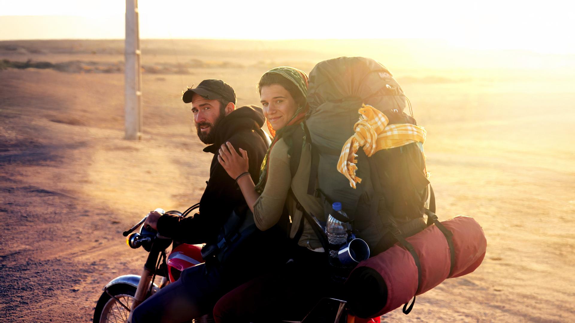 Ein junger Mann mit Bart und Baseballkappe sitzt am Lenker eines Motorrads und schaut in die Kamera. Hinter ihm sitzt eine junge Frau, die sich an seiner Schulter festhält, die Haare mit einem Tuch bedeckt hat und einen vollbepackten Rucksack auf den Schultern trägt. Im Hintergrund ist eine weite Steppenlandschaft unscharf und in einem goldgelben Licht der Abendsonne zu sehen.