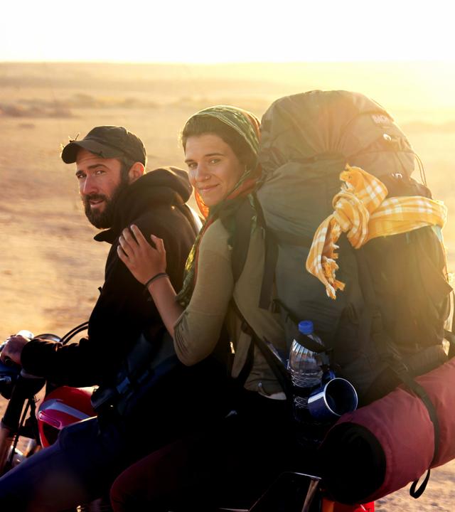Ein junger Mann mit Bart und Baseballkappe sitzt am Lenker eines Motorrads und schaut in die Kamera. Hinter ihm sitzt eine junge Frau, die sich an seiner Schulter festhält, die Haare mit einem Tuch bedeckt hat und einen vollbepackten Rucksack auf den Schultern trägt. Im Hintergrund ist eine weite Steppenlandschaft unscharf und in einem goldgelben Licht der Abendsonne zu sehen.