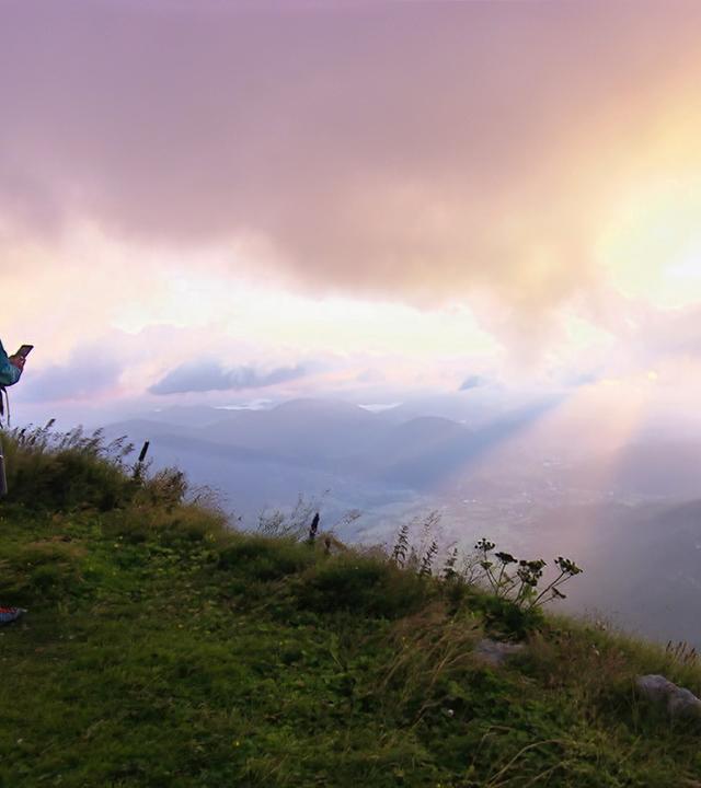Die Darstellung zeigt eine Gruppe von Wanderern an einem Berg, wahrscheinlich in Niederösterreich. Im Vordergrund steht eine Person in Wanderkleidung mit Rucksack, die in die Ferne schaut und möglicherweise ein Smartphone in der Hand hält. Weitere Wanderer stehen daneben und beobachten die Aussicht. Der Hintergrund ist von dramatischen Wolken geprägt, durch die Lichtstrahlen scheinen. Die Landschaft ist hügelig und grün, mit einer Atmosphäre von Weite und Erhabenheit.   Die Aufnahme scheint während der goldenen Stunde gemacht worden zu sein, da eine warme Lichtstimmung herrscht. Die Szenerie vermittelt ein Gefühl der Erkundung und des Abenteuers in der Natur.