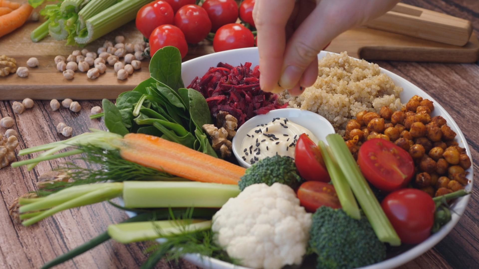 Ein Teller mit Blumenkohl, Broccoli, Sellerie, Möhren, Spinat, Rotkohl, Quinoa, Kichererbsen, Tomaten und einem Dipp: von oben greift eine Hand zu.