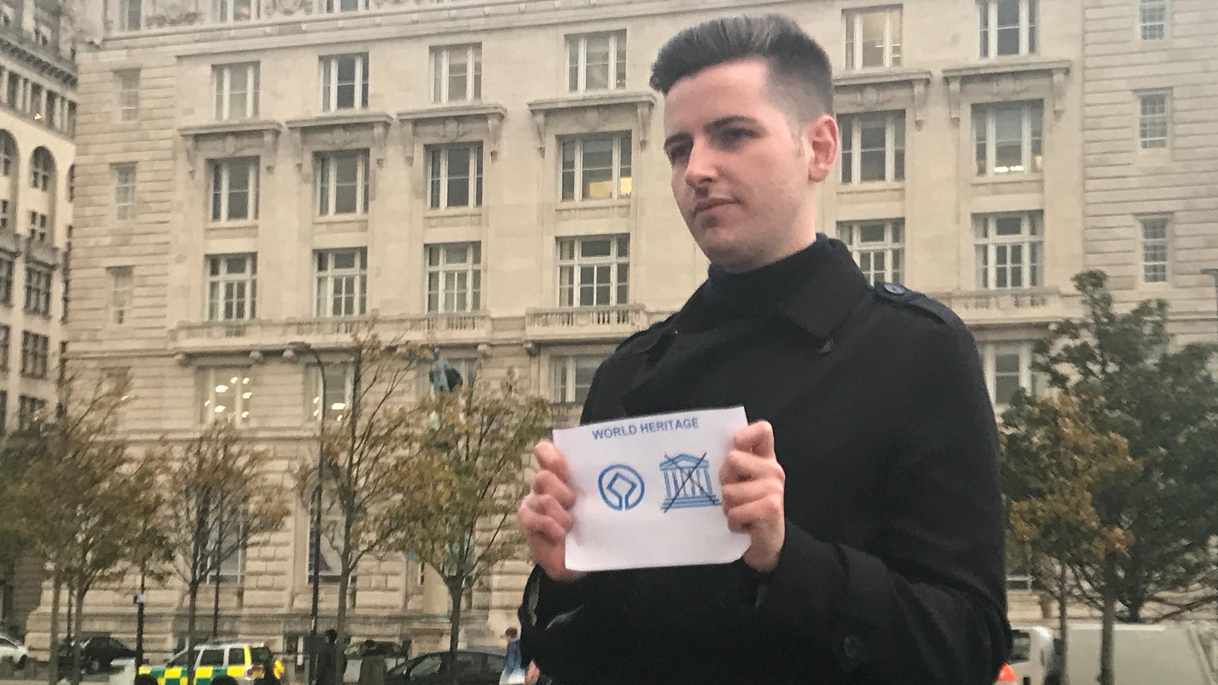 Junger Mann mit durchgestrichenem UNESCO-Label vor Gebäude.