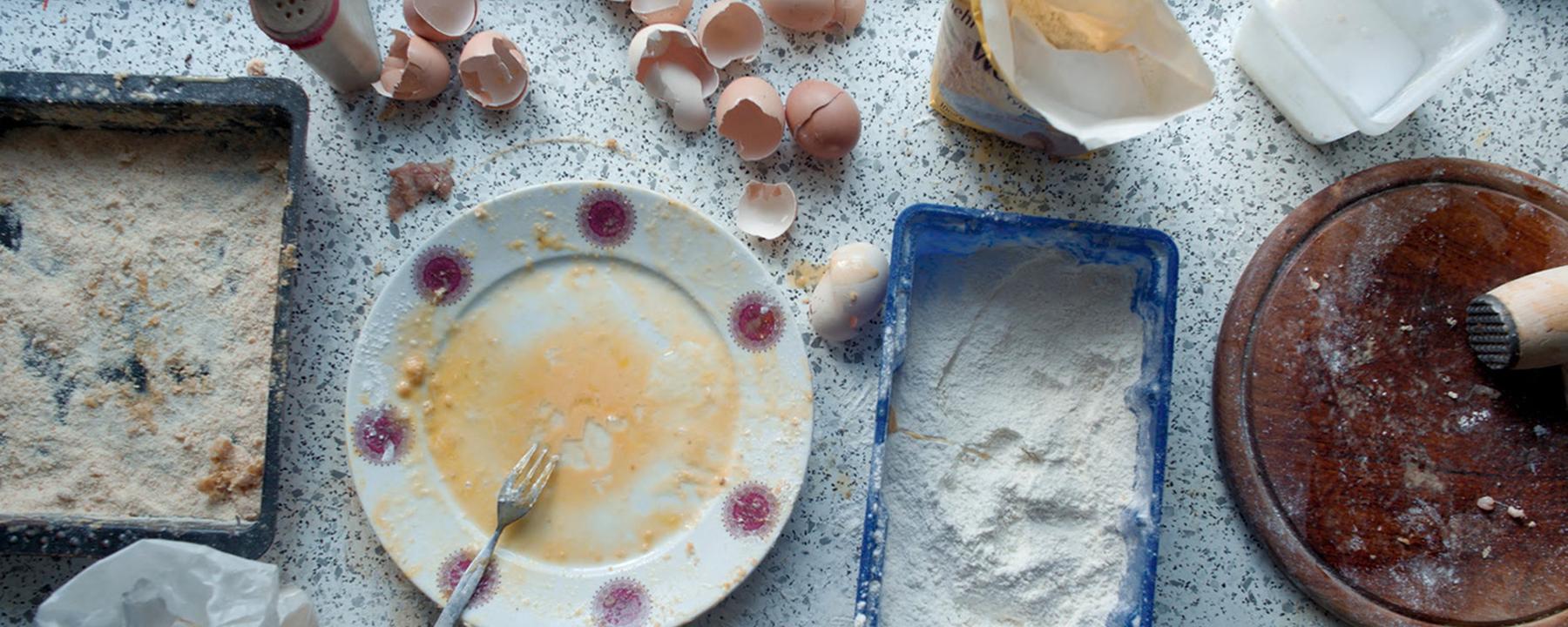 Auf einer Küchenanrichte sieht man den Teil eines panierten Schnitzels, Gefäße mit Mehl, verrührten Eiern und Paniermehl und ein Holzbrett mit einem Fleischklopfer. Daneben liegen mehrere Eierschalen, ein Salzstreuer und Lebensmitteltüten.