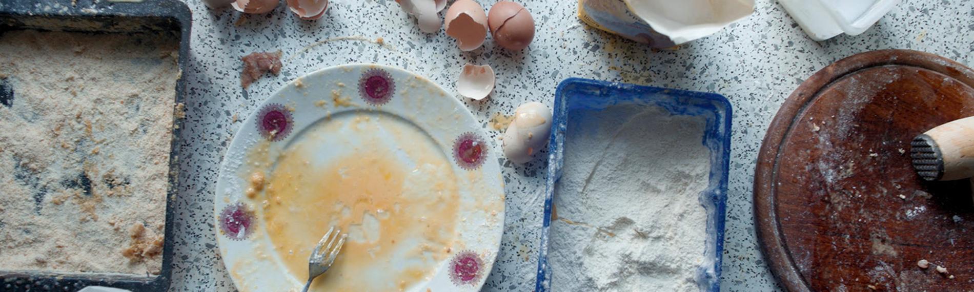 Auf einer Küchenanrichte sieht man den Teil eines panierten Schnitzels, Gefäße mit Mehl, verrührten Eiern und Paniermehl und ein Holzbrett mit einem Fleischklopfer. Daneben liegen mehrere Eierschalen, ein Salzstreuer und Lebensmitteltüten.