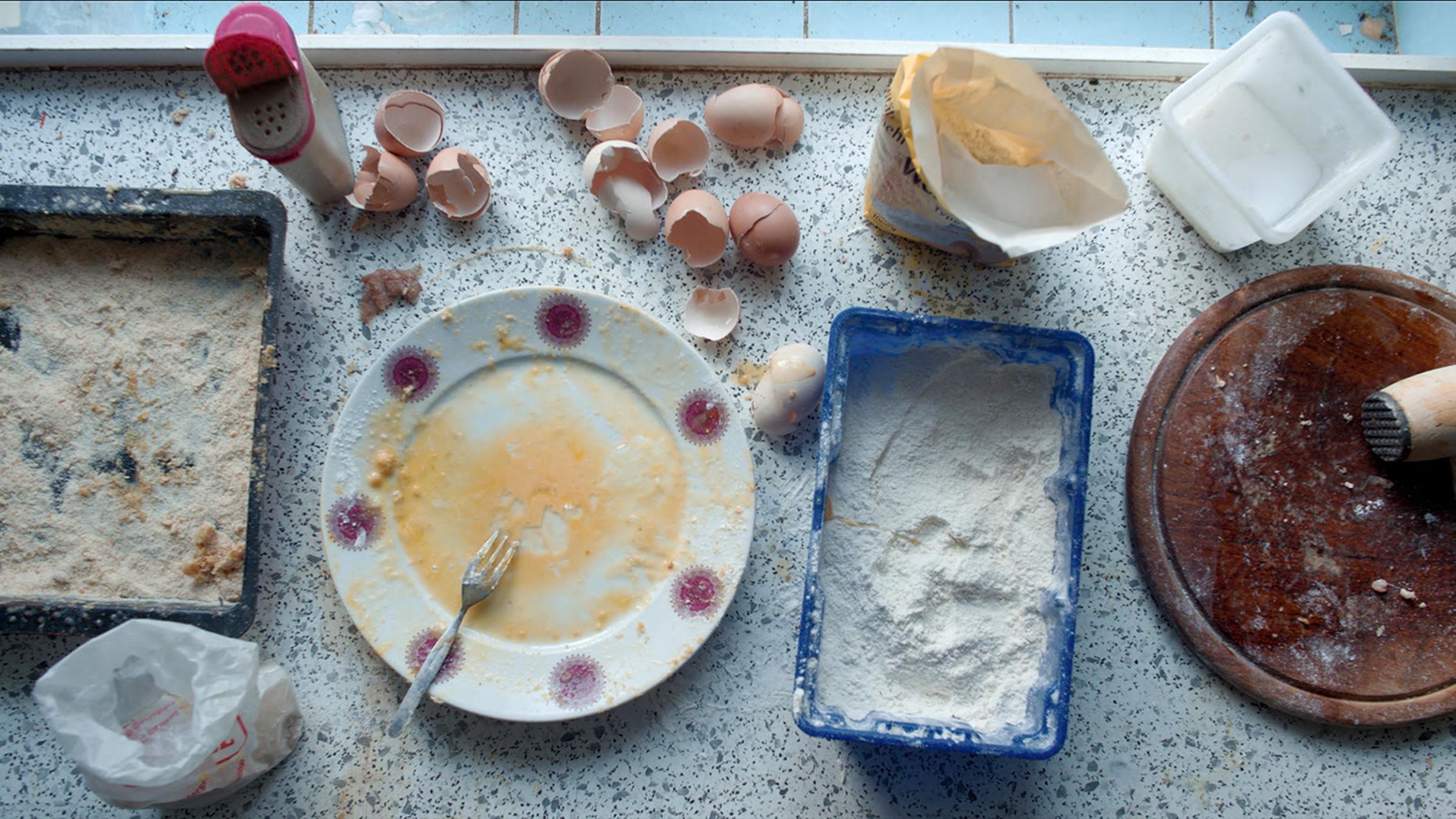 Auf einer Küchenanrichte sieht man den Teil eines panierten Schnitzels, Gefäße mit Mehl, verrührten Eiern und Paniermehl und ein Holzbrett mit einem Fleischklopfer. Daneben liegen mehrere Eierschalen, ein Salzstreuer und Lebensmitteltüten.