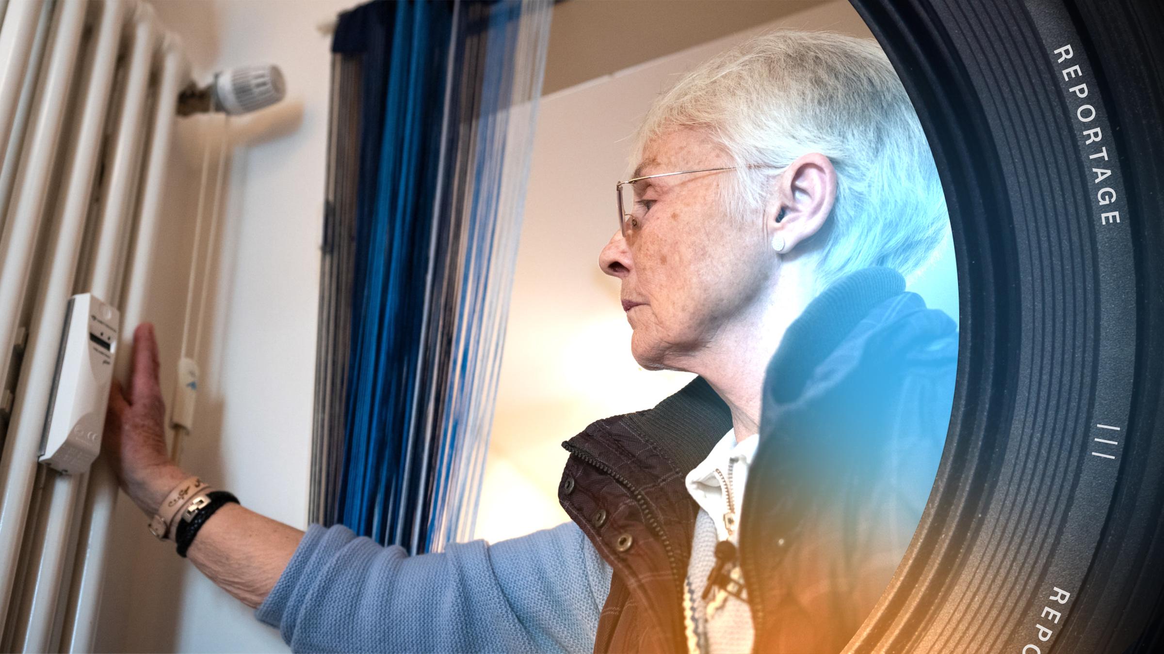 Eine ältere Frau hält eine Hand an eine Heizung und schaut auf ein Messgerät an der Heizung.