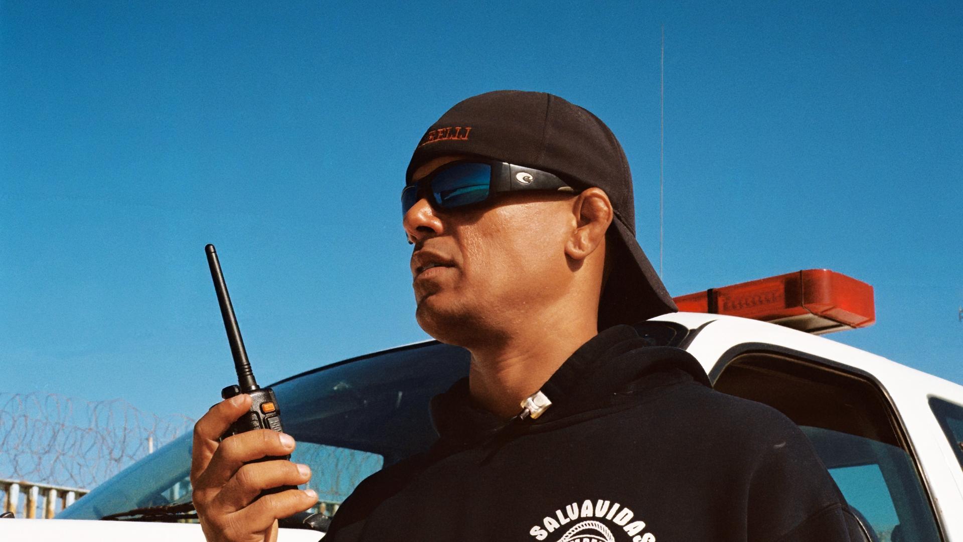 Rettungsschwimmer mit Sonnenbrille, Baseball-Kappe und Funkgerät vor einem Einsatzfahrzeug mit Sirene, im Hintergrund ein Metallzaun mit Stacheldraht.