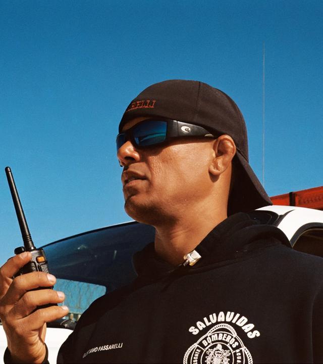 Rettungsschwimmer mit Sonnenbrille, Baseball-Kappe und Funkgerät vor einem Einsatzfahrzeug mit Sirene, im Hintergrund ein Metallzaun mit Stacheldraht.