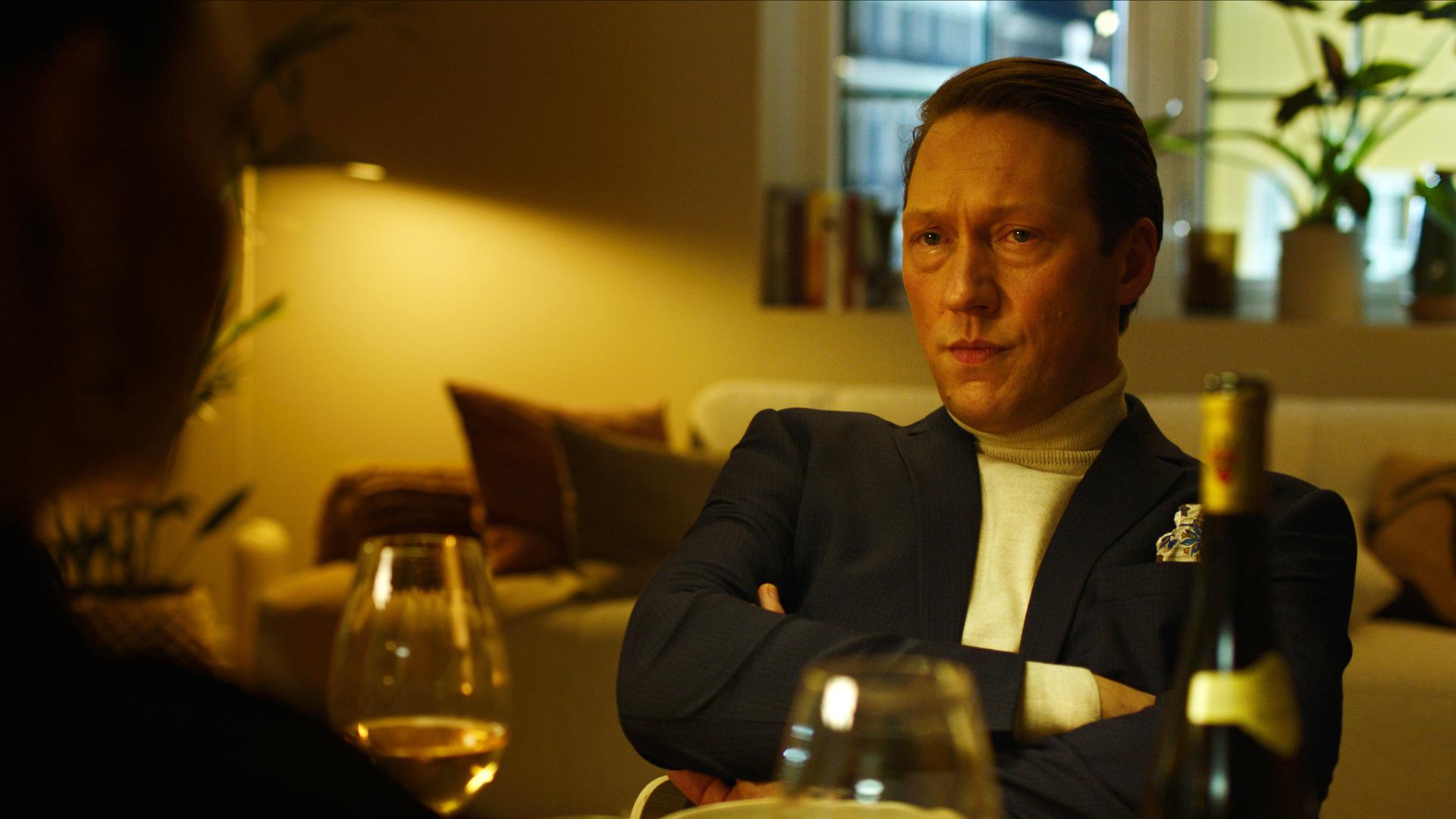 "Exit - Wenn die Welt verschwindet": Adam (Simon J. Berger) sitzt mit verschränkten Armen am Esstisch und schaut sie ernst an.