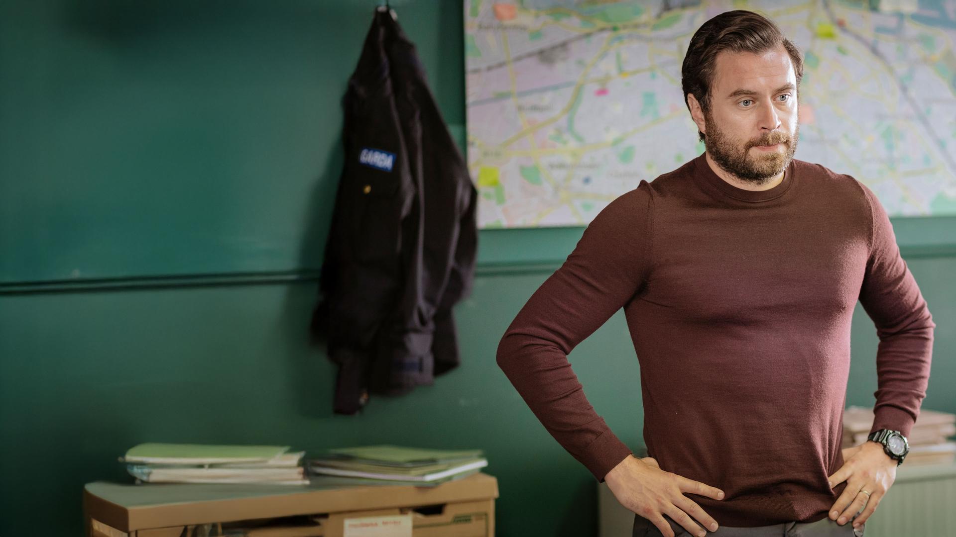 "Harry Wild - Mörderjagd in Dublin: Wenn Happy nicht happy ist, ist niemand happy": Charlie Wild (Kevin J. Ryan) steht in einem Büro, seine Arme in die Hüften gestützt. Er schaut ernst.