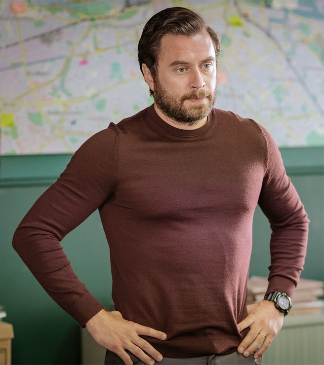 "Harry Wild - Mörderjagd in Dublin: Wenn Happy nicht happy ist, ist niemand happy": Charlie Wild (Kevin J. Ryan) steht in einem Büro, seine Arme in die Hüften gestützt. Er schaut ernst.