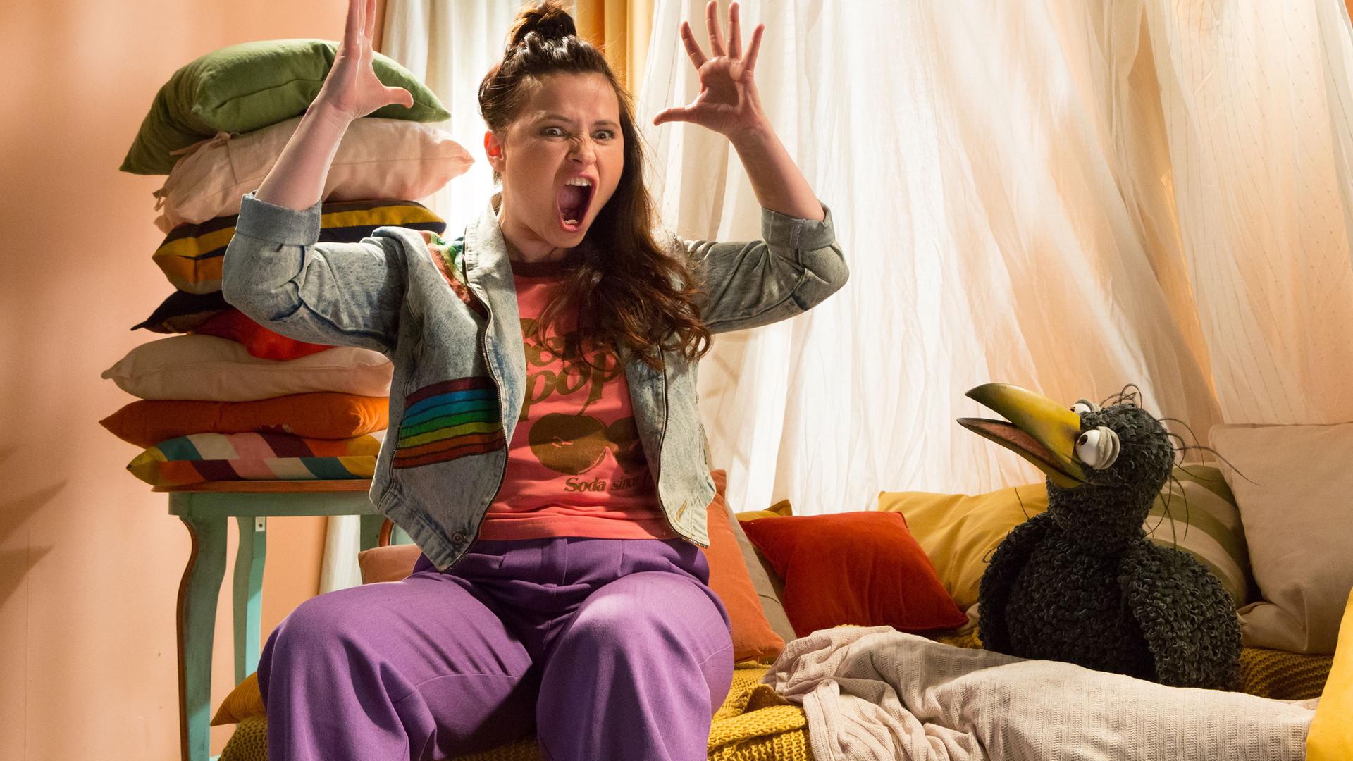 Rudi hockt in der Kuschelecke und schaut neugierig auf Anna, die neben ihm sitzt. Sie hebt die Arme und reißt wütend Mund und Augen auf.