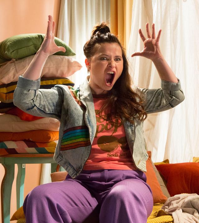 Rudi hockt in der Kuschelecke und schaut neugierig auf Anna, die neben ihm sitzt. Sie hebt die Arme und reißt wütend Mund und Augen auf.